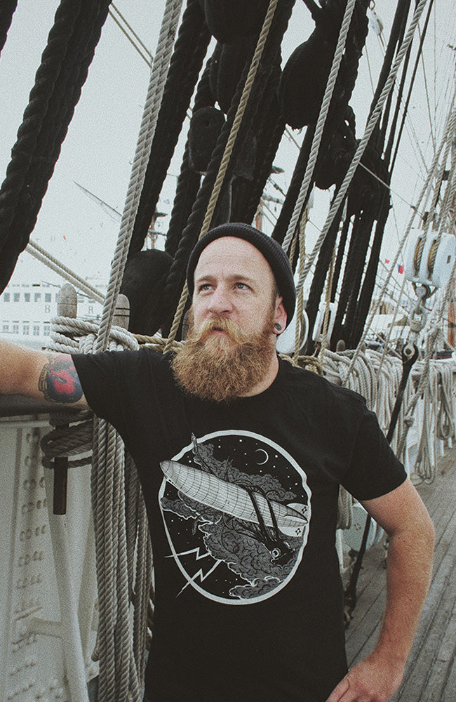 A person with a beard wearing a black t-shirt standing on a boat with nautical ropes in the background. The t-shirt features a graphic of a storm with a giant squid and a zeppelin.
