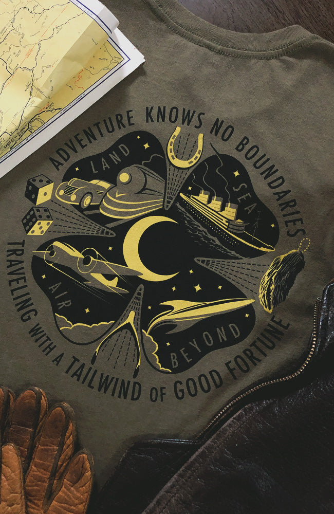A green t-shirt laid out flat with a graphic design featuring text and symbols related to travel and luck, including a four-leaf clover, crescent moon, lucky horseshoe, lucky rabbit's foot, two dice, vintage car, train, art deco ship, airplane and rocket into the stars. Printed text of "Adventure knows no boundaries, traveling with a tailwind of good fortune"