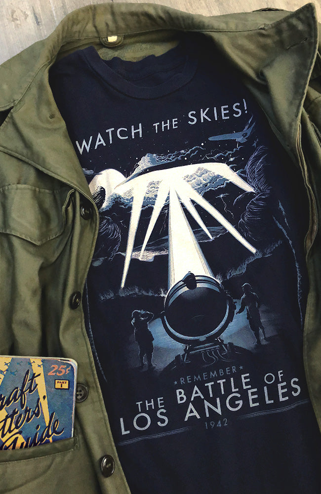 Navy color unisex t-shirt with a graphic print of anti-aircraft guns firing and the text 'THE BATTLE OF LOS ANGELES 1942' on it, displayed under a jacket.