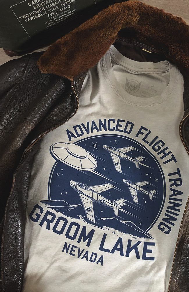 Gray commemorative t-shirt with 'Advanced Flight Training Groom Lake Nevada' text and vintage airplane graphics, displayed alongside a brown leather jacket.
