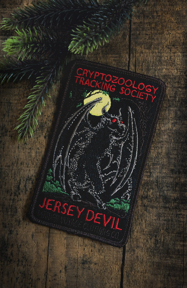 A patch featuring a black Jersey Devil creature with red eyes, surrounded by text 'CRYPTOZOLOGY TRACKING SOCIETY JERSEY DEVIL Maiden Voyage Clothing Co.', placed on a wooden surface with greenery in the background.