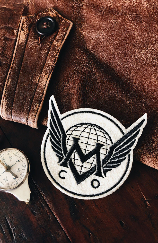 A vintage inspired iron-on Maiden Voyage Co. logo patch with ivory wings placed on a brown leather surface next to a white compass.