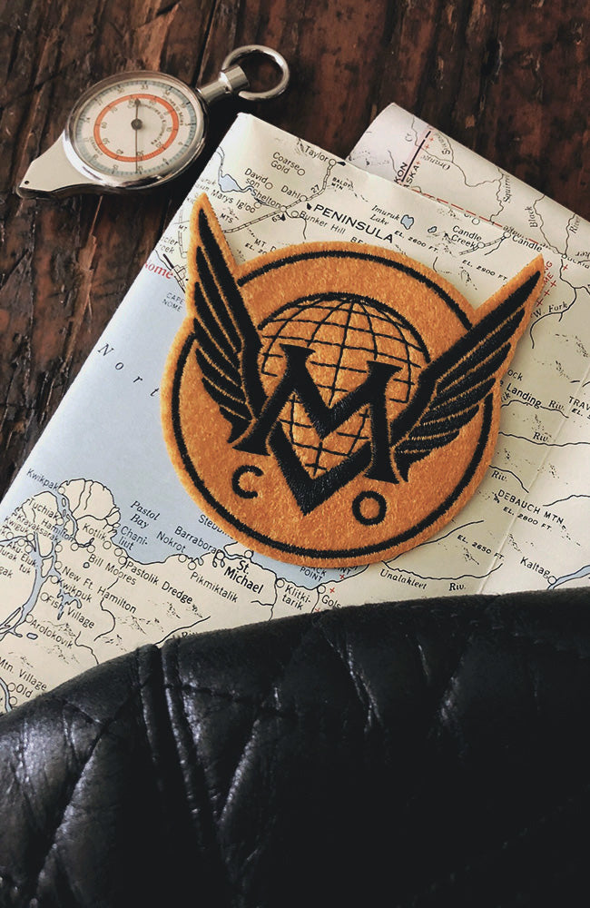 An iron-on patch with a vintage style aviation themed Maiden Voyage Co.  logo placed on a map, next to a compass and a leather object.
