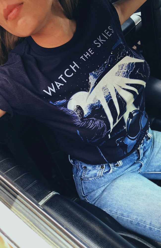 A person wearing a navy-colored unisex t-shirt with a graphic print of anti-aircraft guns and text 'WATCH THE SKIES'.