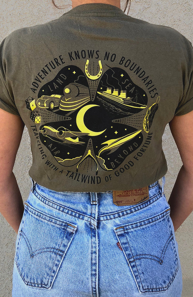 A green t-shirt laid out flat with a graphic design featuring text and symbols related to travel and luck, including a four-leaf clover, crescent moon, lucky horseshoe, lucky rabbit's foot, two dice, vintage car, train, art deco ship, airplane and rocket into the stars. Printed text of "Adventure knows no boundaries, traveling with a tailwind of good fortune"